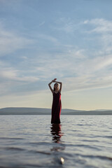 Beautiful woman in light red dress stands in water of lake sea at sunset. Reflection of the girl and sky in water
