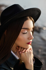 Portrait young woman at sunset. Fashion woman in black round hat and leather raincoat near lake pond. Beautiful makeup and bright red lipstick on lips
