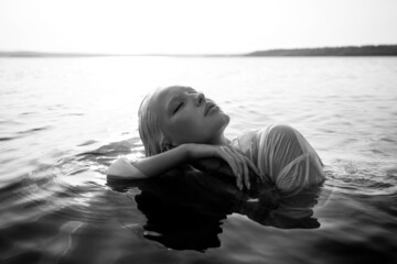 Fototapeta premium Beautiful blonde woman short hair lies in water on sea shore, pond. Art portrait of a relaxing woman in the water on shore
