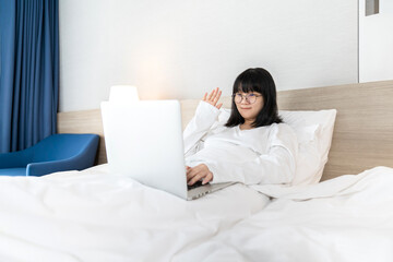 Glasses Asian young pretty teenager rise hand up hi and hello to laptop notebook for meeting and video conference on the bed in Work From Home period.