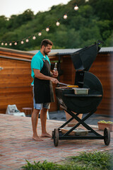Young adult man wearing apron, holding bottle of beer and barbecuing outdoors - obrazy, fototapety, plakaty