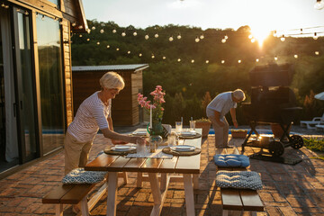 Senior couple preparing family gathering in the backyard. Woman setting table while man barbecuing
