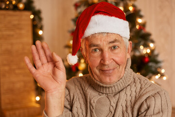 Indoor shot of positive optimistic mature man wearing warm sweater and santa claus hat, looking at camera and waving hand, posing in festive living room at home near xmas tree.