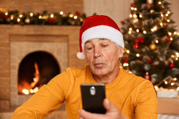 Concentrated attentive mature man wearing orange sweater and santa claus hat using mobile phone while posing at home in decorated living room, looking at cell phone display.