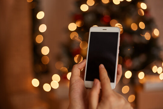 Image of smart phone in woman's hands with blurred Christmas tree with garland lights on background, mobile phone with blank screen for advertisement or promotional text.
