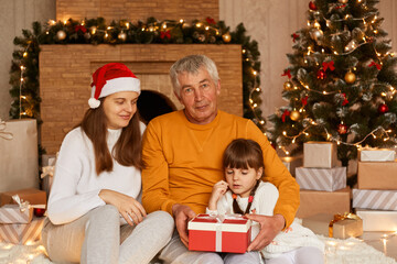 Indoor shot of happy family giving Christmas present to little cute dark haired girl in red wrapped gift box, posing in decorated living room near xmas tree and fireplace.