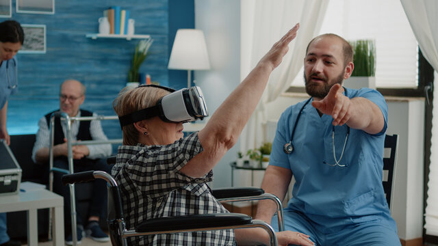 Disabled old woman in wheelchair using vr glasses in nursing home while nurse explaining virtual reality for healthcare and recovery treatment. Patient with physical issues and technology