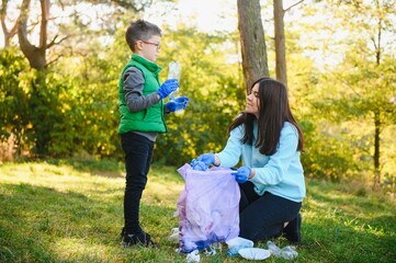 Fototapeta premium Smiling boy picking up trash in the park with his mother. Volunteer concept.