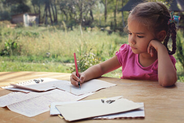 A little girl writing a letter with ink pen, old envelopes, ink pen and set of replacement pens on...