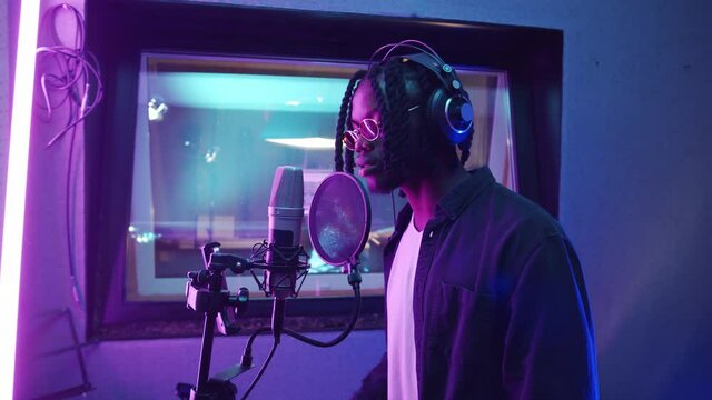 Close up shot of young African American rapper with dreadlocks singing in microphone in recording studio with purple light