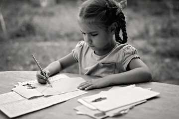 A little girl writing a letter with ink pen, old envelopes, ink pen and set of replacement pens on the aged wooden table, retro and vintage concept, black and white toning