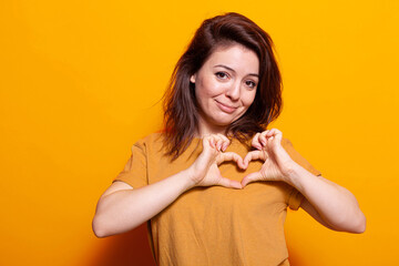 Caucasian woman showing heart shape with fingers at camera. Portrait of romantic person doing love sign and symbol with hands, showing affection and emotion over orange background.