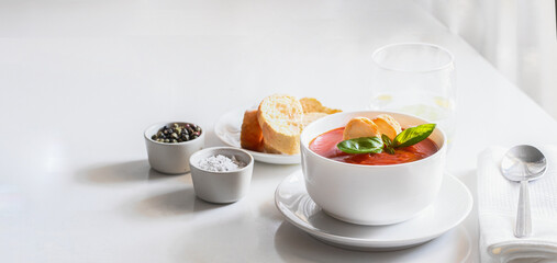 Tomato soup plate decorated with bread crumbs, fresh basil  and herbs on the restaurant table with water glass ,portion of bread  and indivudual spices bowls.Banner with copy space