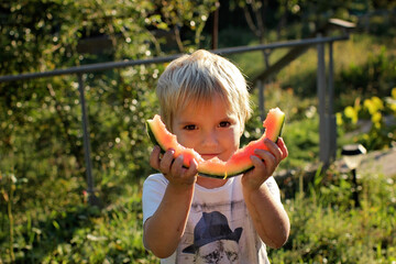 A three year blond boy eating a tasty watermelon in the garden of country house at summer, happy...