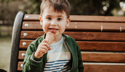 Close-up portrait of caucasian boy holding ice cream in his hand on the bench in a park, looking and smiling at camera messed up and dirty