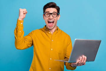Photo of astonished crazy guy hold laptop scream raise fist wear specs yellow shirt isolated blue...