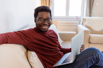 Portrait of smiling African American man in glasses sit at sofa in office working on laptop, happy biracial male worker look at camera posing, busy using modern computer gadget at workplace