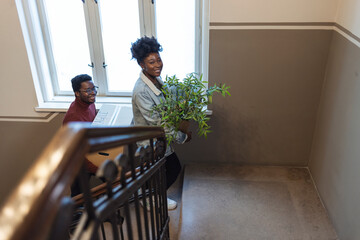 Young couple carrying big cardboard box into new home. Moving house. Couple carrying boxes up the staircase while moving into a new building. Young happy black couple relocating into new home.