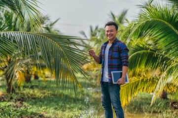portrait of middle aged man hold tablet, standing  and looking at camera, outdoors in coconut farm. Smart farming and digital agriculture concept