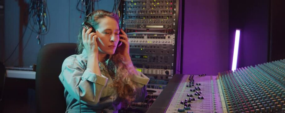 Cheerful woman using mixing console, then taking off headphones and posing for camera with smile while working in recording studio