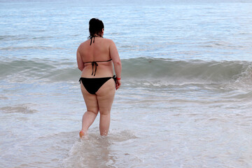 Fototapeta na wymiar Overweight woman in black swimsuit going to swim in a sea water. Vacation on a beach, overeating and weight loss concept