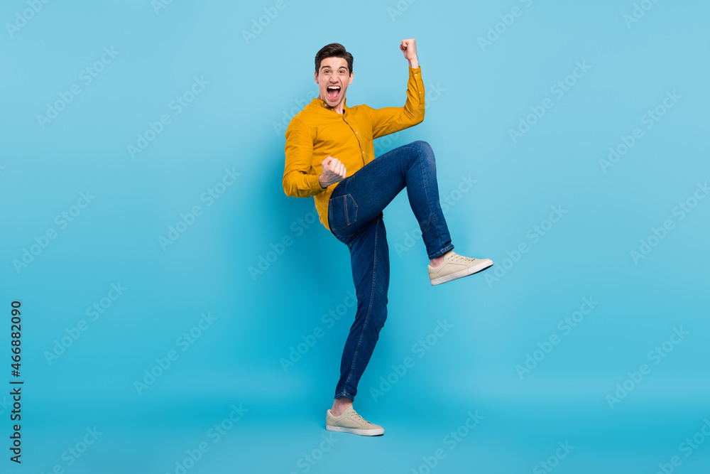 Poster Photo of pretty lucky young guy dressed yellow smiling walking rising fists isolated blue color background