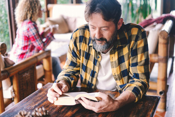 Adult man reading a book at home sitting on the wooden table with wife woman in background. Happy...