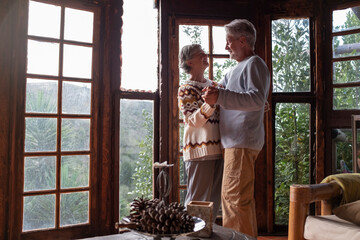 Happy senior couple in love dance at home agains a big windows view with nature woods outside....