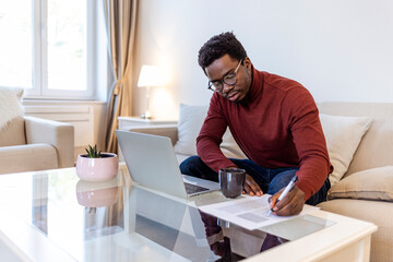 Fototapeta na wymiar African-American male student or worker in glasses is watching online lectures or webinars and writing notes in a notebook. Concentrated dark skinned copywriter in eyewear typing text of publication 
