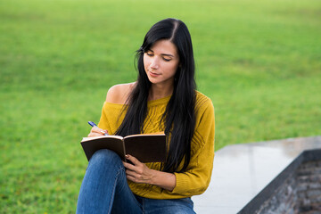 Obraz premium A beautiful and attractive Caucasian brunette girl in a yellow sweater writing in a notebook while sitting in the park.