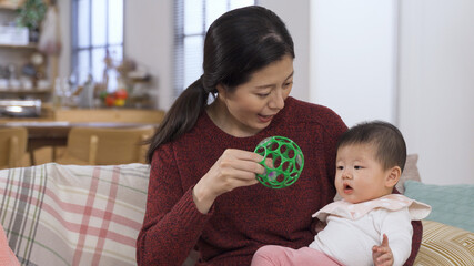 happy asian mother is playing with her lovely baby daughter in arms by shaking a toy ball to her and putting it her hands on sofa at home.