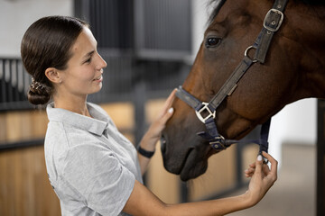 Side view of female horseman taking care of her brown Thoroughbred horse in stable. Concept of animal care. Rural rest and leisure. Idea of green tourism. Young european woman wearing uniform
