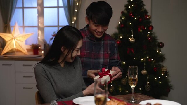 hide and seek time for presents. Loving asian husband closing wife eyes with hand and prepared Christmas surprise. cheerful girl laughing getting gift during xmas dinner time