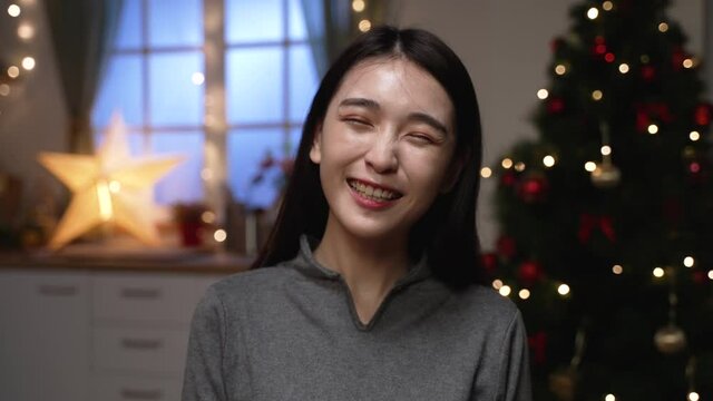 Headshot smiling Asian woman greeting friends looking at camera while making video call on Christmas tree background. happy female blogger recording vlog. young girl relax talking chatting online
