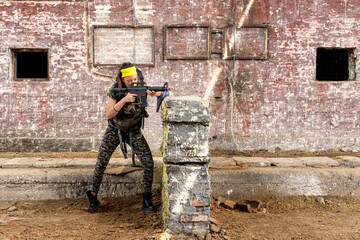 Strong young woman hiding behind wall with backpack and riffle on hans in old industrial place