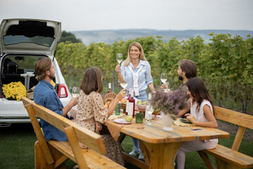 Joyful european friends drinking and toasting with local wine during picnic near vineyards in countryside. Young men and women spend time together. Concept of winemaking. Friendship and leisure.
