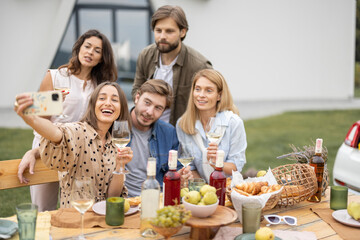 Pleased european friends taking selfie on mobile phone during friendly picnic. Young men and women drinking wine from glasses. People enjoying time together. Concept of friendship. Idea of leisure