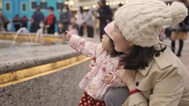 closeup view of happy asian mother and curious baby girl having fun looking at beautiful water fountain in downtown area during Christmas season