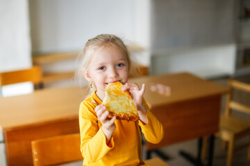 Cute caucasian girl child blonde eating a slice of pizza with cheese, a snack at school, junk food and fast food at children