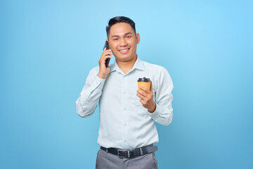 Fototapeta premium Smiling young handsome businessman talking on smartphone and hold a cup of coffee on blue background