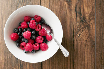 Raspberry and blueberry in a white bowl on a wooden table. Healthy fruit mix. Produce product. Healthy diet choice.