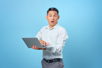 Portrait of shocked young handsome businessman holding and using a laptop on blue background