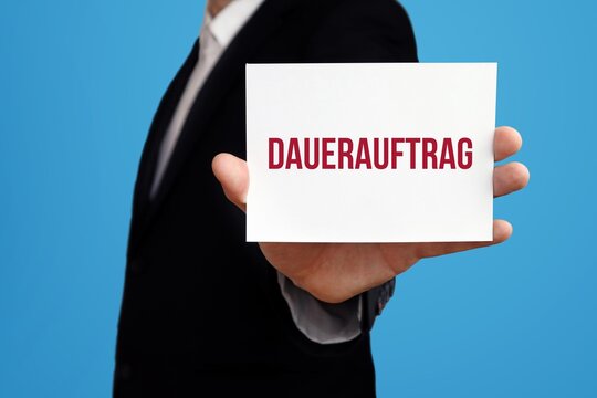 Dauerauftrag &Uuml;berweisung. Gesch&auml;ftsmann im Anzug zeigt Karte mit Text. Mann isoliert vor Hintergrund (blau)