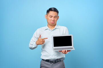 Sad young handsome businessman pointing at a laptop with a blank screen on blue background