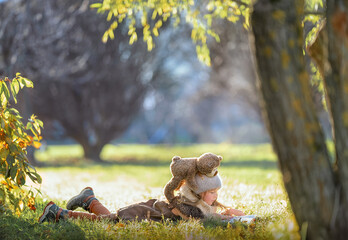 Obraz premium Little Girl with her teddy bear friend reads a book of fairy tales in nature, lying on the lawn in the beautiful light of the sun
