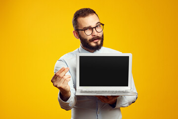 Bearded man in eyeglasses and blue holding new modern laptop device while showing money gesture,...