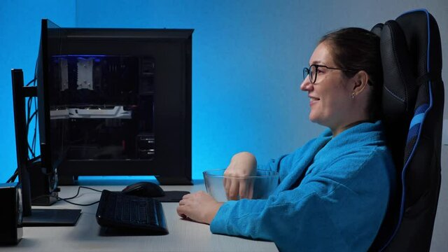 Young brunette woman in glasses wearing blue bathrobe smiles watching stream with interest on large computer screen eating popcorn and sitting in dark room.