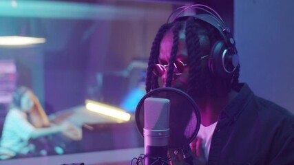Young African American rapper in headphones singing in microphone while recording music in audio production studio