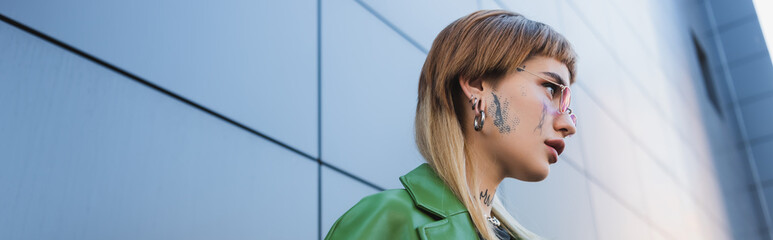 low angle view of young woman with tattoo and piercing near grey wall outdoors, banner