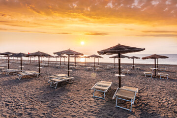 scenic view at nice sunset or sunrise beach with rows of umbrellas and chaise lounges with blue sea and amazing cloudy sky with sun glow on the background